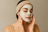 A serene woman with a beige headband gently touches her face while wearing a creamy white facial mask, highlighting relaxation and skincare in a bright, minimal setting.